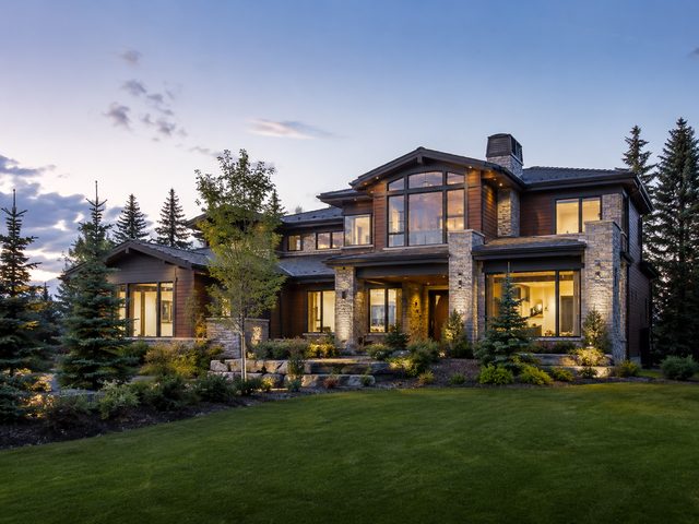 A large contemporary home lit at dusk behind mature spruce trees on a wide Calgary lot.